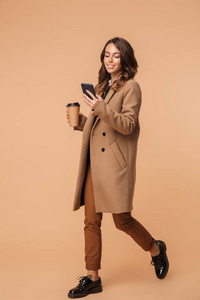 这位20多岁的迷人女人穿着外套，拿着手机和外卖咖啡，站在米色背景上孤立无援。