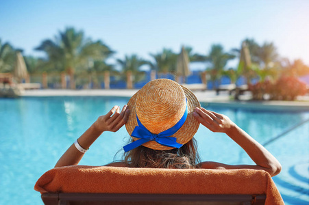 坐在阳光躺椅上的年轻美女在酒店的游泳池戴着帽子