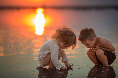 日落时，两个小孩子在海滩上玩耍。孩子们在水里玩。水的反射