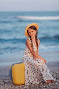 一个小女孩坐在海滩上的黄色手提箱上。戴帽子的女孩的肖像。复古风格