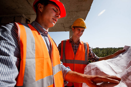建筑师和结构工程师穿着衬衫橙色工作服和头盔, 在在建建筑内的建筑工地上探索施工文件