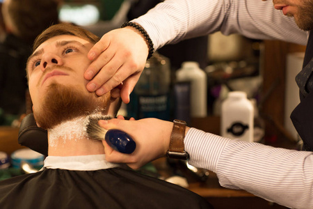 在理发店的专业理发师那里，一个英俊的留着胡须的年轻男子在他的客户服务剃须风格时尚男性化的颈部涂上剃须膏。