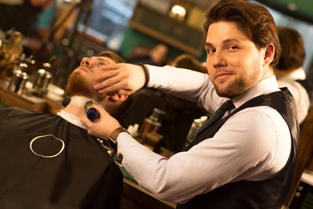 开朗英俊的胡须专业理发师微笑着对着相机，同时剃掉他的男客户在理发店复印空间工作成功自信专业理发职业工作。