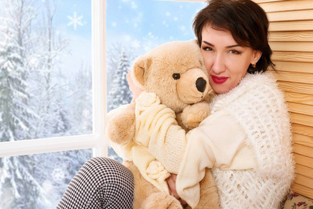 女人坐在窗台上拿着熊玩具。 窗外美丽的景色，阳光明媚的冬天，森林和雪。