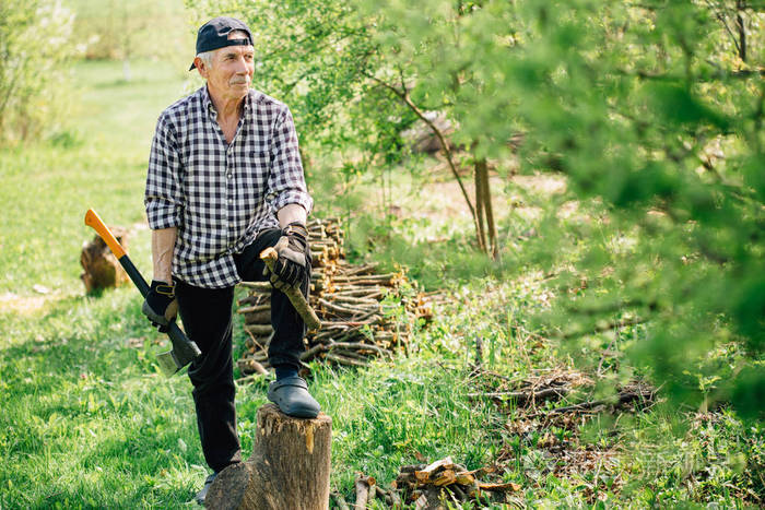 戴棒球帽的老人用斧头砍木头. 年长的植树者在花园里工作.