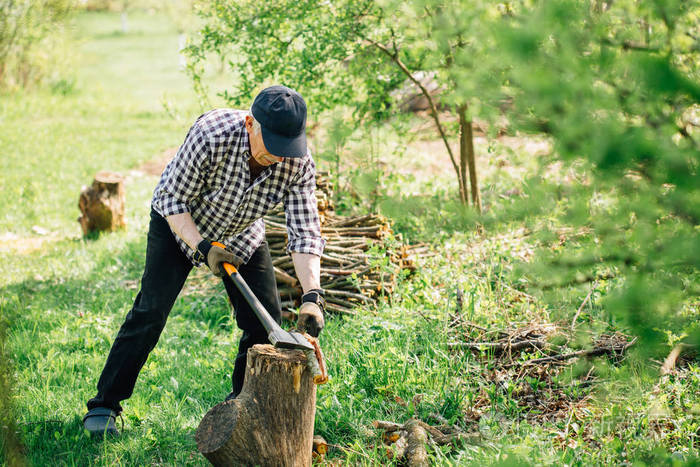 戴棒球帽的老人用斧头砍木头一个在花园里工作的老人