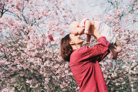 慈爱的母亲和女婴在背景上绽放粉红色的苹果树花在春天。 美丽的女人和女儿在户外的花园花中。 植物园自然的家庭