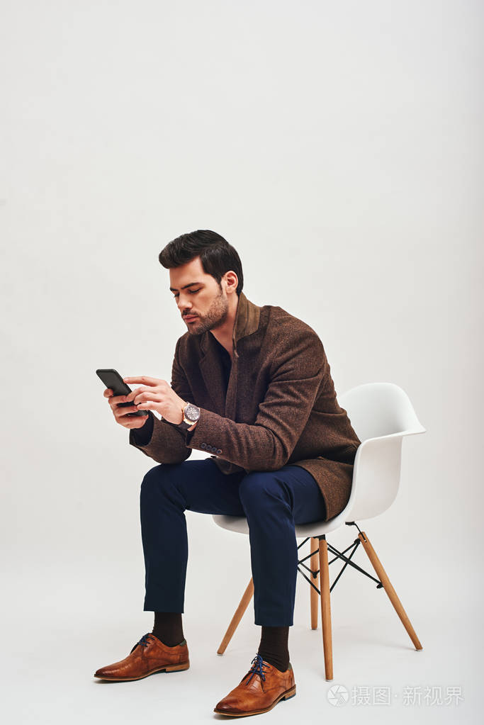 时尚的黑发男子坐在椅子上, 在他的手机里搜索孤立的白色背景
