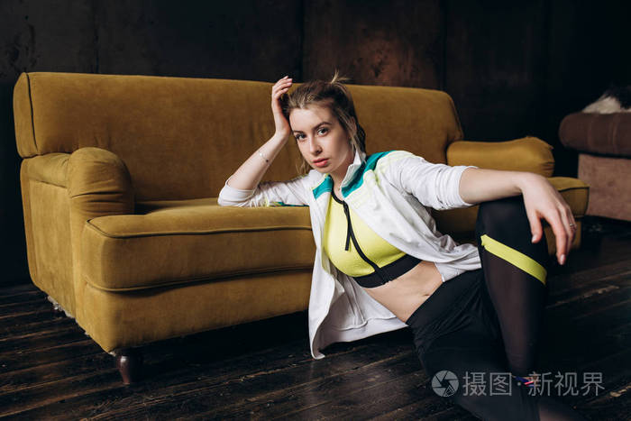 一位身穿运动服,坐在地板上的黄色沙发旁摆姿势的年轻健身女子的全身