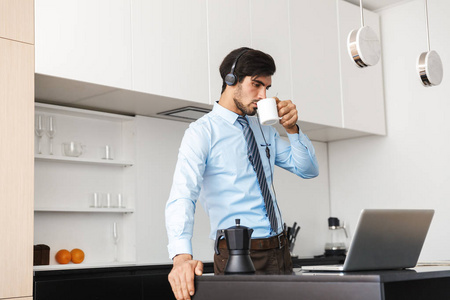 自信的年轻商人在厨房里工作，用笔记本电脑通过耳机连接，喝咖啡