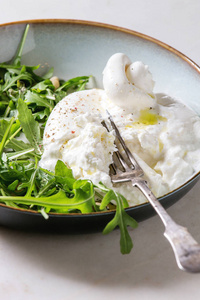 切片意大利波拉塔奶酪新鲜阿鲁古拉沙拉松坚果和橄榄油在白色大理石桌子上的白色陶瓷板。 近点