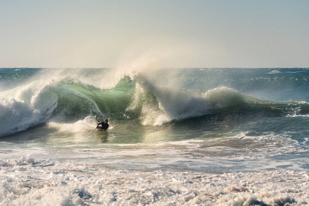 人冲浪是一个巨大的波浪，它以大量的能量和力量打破，阳光在海的绿松石表面反射金色，在巨大的泡沫风中升起羽状的水