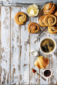各种自制的泡芙糕点面包，肉桂卷和牛角面包，与咖啡杯果酱黄油作为早餐，白色木板背景。 平躺空间
