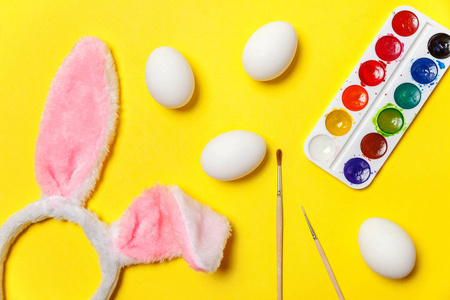 快乐复活节的概念。 为假期做准备。 装饰鸡蛋，彩色油漆和兔子耳朵，毛茸茸的服装玩具，孤立在时尚的黄色背景上。 简单的极简主义，平