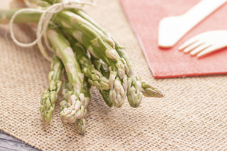 新鲜的绿芦笋放在老的餐桌上，用环保的木刀和叉子放在红色的餐巾纸上。健康的天然食物。