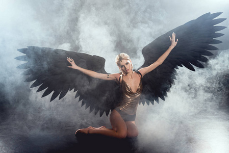 美丽的女人，有着黑色的天使翅膀，伸出双手，坐在黑暗的烟雾背景上摆姿势
