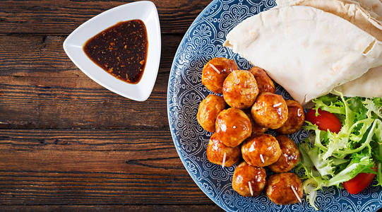 肉丸在糖和酸釉的盘子上，皮塔面包和蔬菜在摩洛哥风格的木桌上。 塔帕斯。 趋势食品。 顶部视图