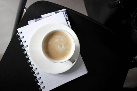 卡布奇诺放在黑酒吧椅上的笔记本上。一杯新煮的咖啡，加上美味的牛奶泡沫。最低限度的室内设计。