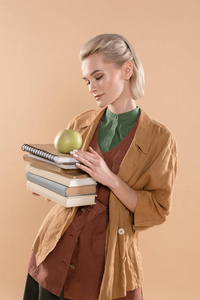 美丽的女孩拿着书和绿苹果，站在生态服装中，被隔离在米色中