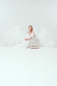 美丽的女人，穿着天使服装，翅膀坐着，摆姿势，鸟笼被隔离在白色上