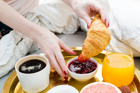 早餐吃牛角面包的女孩在床上酒店服务。 咖啡果酱牛角面包橙汁葡萄柚荔枝。 侧视灯光背景关闭