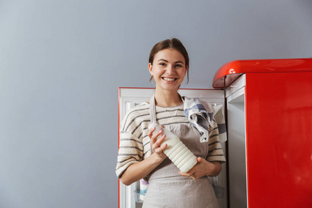 幸福的年轻女子站在家里的厨房里，从冰箱里拿出一瓶牛奶