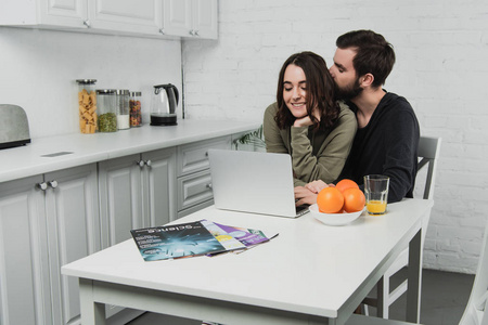 帅哥抱着女人坐在桌子上，拿着橘子，在厨房里用笔记本电脑