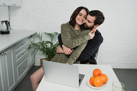 在厨房吃早餐时，一对漂亮的幸福夫妇抱在一起，坐在桌上，手提电脑和橘子