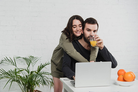 美丽的微笑的年轻女子拥抱男人，用笔记本电脑，在厨房早餐时喝橙汁