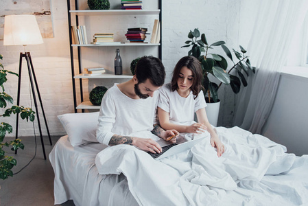 一对年轻漂亮的夫妇，穿着睡衣躺在床上，用笔记本电脑