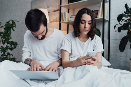 一对年轻漂亮的夫妇穿着睡衣躺在床上，一边用笔记本电脑和智能手机