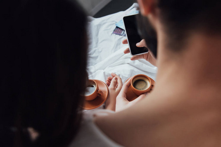 背面视图夫妇与咖啡杯使用智能手机与空白屏幕在家里。