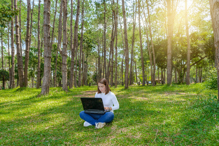 美丽的年轻女子坐在公园的绿草上的肖像，在夏天的一天，在使用笔记本电脑时交叉着双腿。 背景是松林景观