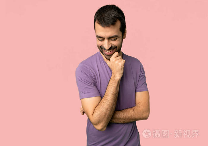 英俊的男人低头看着,手放在孤立的粉红色背景上的下巴上