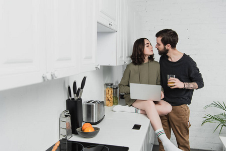 一对漂亮的夫妇，带着橙汁和笔记本电脑，在厨房的早餐期间互相看着对方