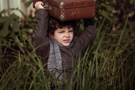 一个穿着外套的小男孩偷偷地穿过高高的草地，头上戴着一个手提箱。 小旅行者。 旅游业。 古董。 从家里逃走。