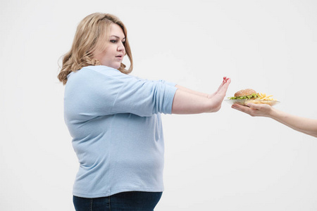 一个年轻的华丽的胖女人穿着休闲的蓝色衣服在白色的背景下拒绝从快餐提供给她, 一个汉堡包和炸薯条。饮食和适当的营养