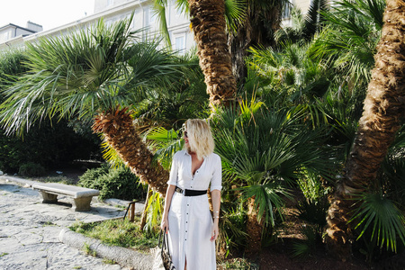 美丽优雅时尚的高加索女人带着太阳镜拿着她的手提包，在棕榈树面前摆姿势。