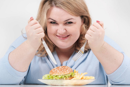 年轻的弯曲脂肪妇女穿着休闲蓝色的衣服, 在白色的背景下, 在桌子上急切地吃着快餐汉堡包和薯条。饮食和适当的营养
