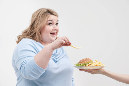 年轻的弯曲脂肪女人穿着休闲的蓝色衣服, 穿着白色的背景, 偷偷从一盘汉堡包和炸薯条上下来。饮食和适当的营养
