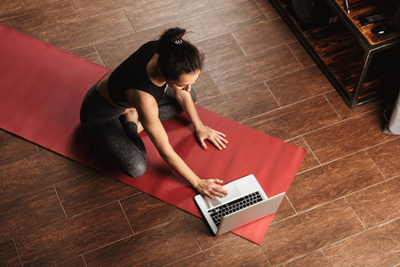 漂亮健康的女人在家里坐在健身垫上，用笔记本电脑做瑜伽练习，伸展