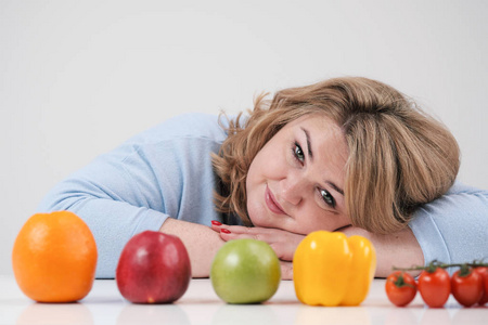 年轻的郁郁葱葱的胖女人穿着休闲的蓝色衣服, 在白色的背景下在桌子上, 在她面前排起了一排的蔬菜和水果。饮食和适当的营养