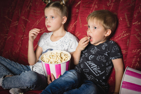 一个女孩和一个男孩在黑暗中的红色大沙发上在家看电影, 吃爆米花照片