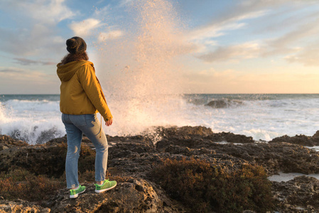 年轻的女人站在岩石上，在冬天的海洋附近享受日落。 冬天去欧洲旅游