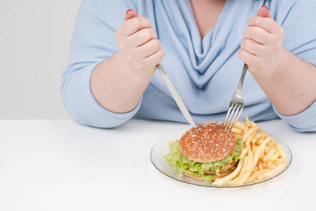 年轻的弯曲脂肪妇女穿着休闲蓝色的衣服, 在白色的背景下, 在桌子上急切地吃着快餐汉堡包和薯条。饮食和适当的营养