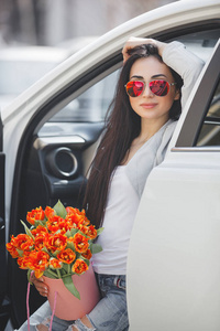 年轻迷人的女人在车里带着礼物盒礼物和鲜花。 春天美丽的女士，带着一束郁金香。 女在汽车里。