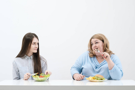 苗条的女孩吃健康的食物, 胖女人吃有害的快餐。在白色背景下, 饮食和适当营养的主题