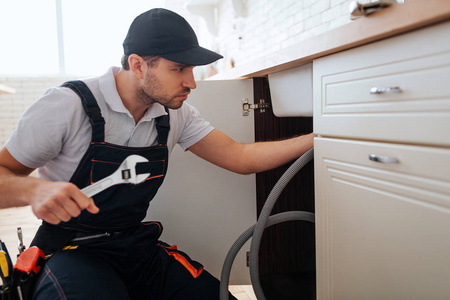 他站在厨房里, 望着水槽下面.管道工软管照片