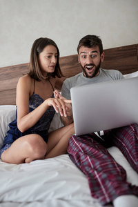 美丽的年轻微笑的夫妇在早上享受。坐在床上拿着笔记本电脑上网