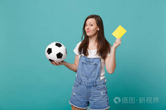 困惑的年轻女子足球球迷支持队与足球黄牌建议球员退休从球场孤立的蓝墙绿松石背景。 家庭休闲的概念